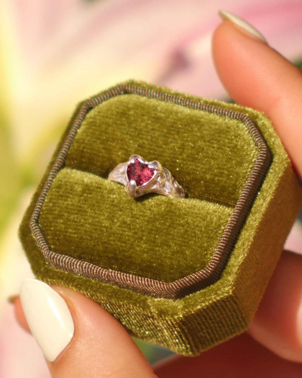 Hier sehen wir eine Nahaufname von folgendem Bild: Eine Hand hält eine Samt grüne Ringbox in der sich ein silberner RIng befindet. In der Mitte des Echtsilber Rings befindet sich ein dunkelrotes Granatherz als Edelstein, dieser ist hangefasst mit 3 Krabbenfassungen. Im huntergrund sieht man verschwommen eine rosane Lilie.